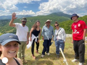 Six archeological team members standing atop a hill fort with green mountains and blue cloudy skies in the background.